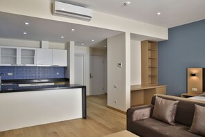 A modern kitchen with white cabinets, a black countertop, and a blue backsplash. There is a brown sofa and a small table with a lamp in the living area.