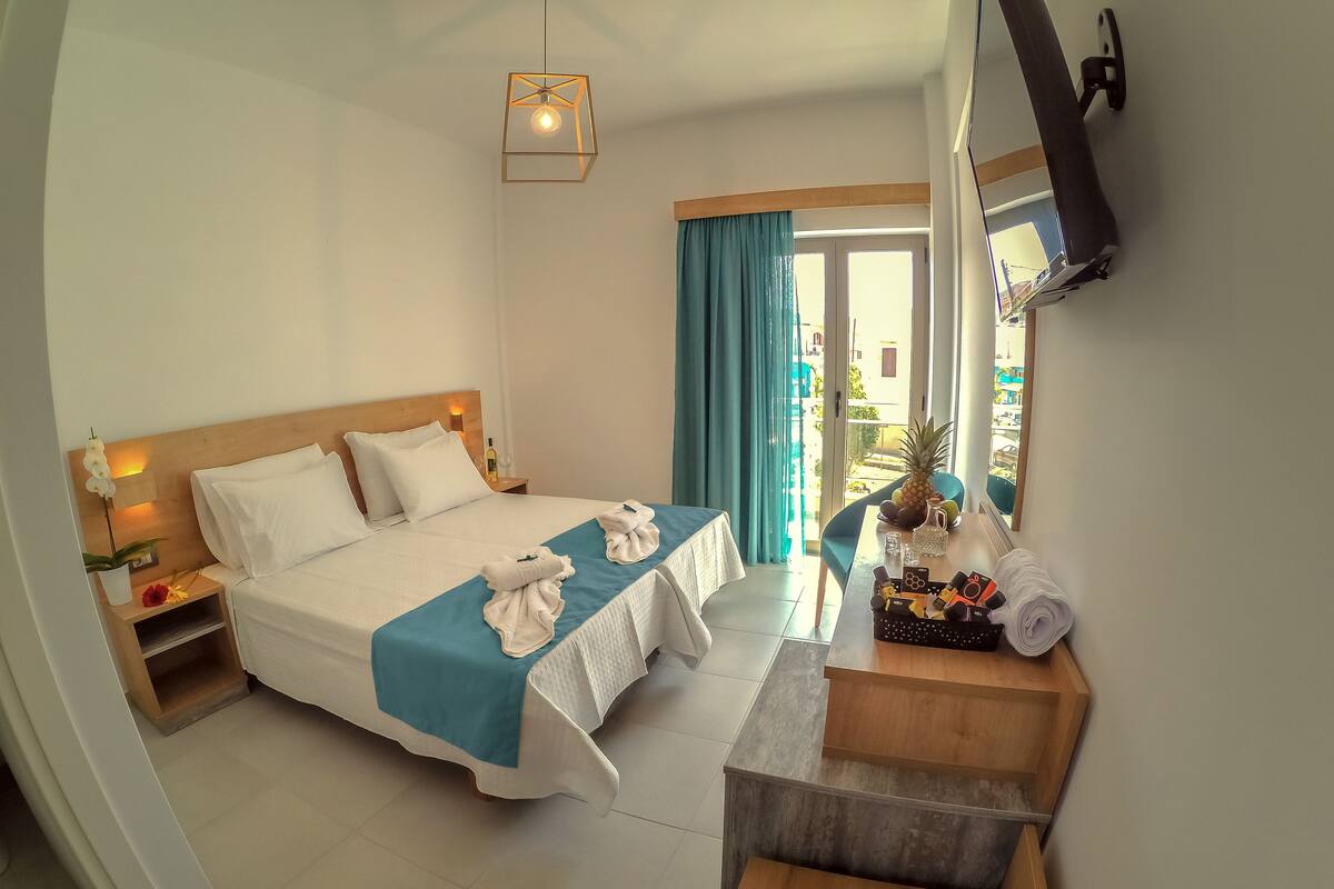 A hotel room with a bed, a desk, a small table with a pineapple and a pitcher, and a view of the outside.