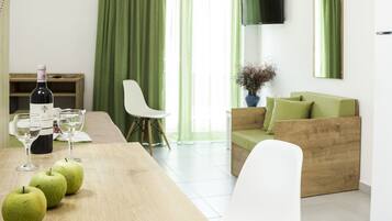 Una sala moderna con una mesa de madera, manzanas verdes, copas de vino, una botella de vino, un sofá verde y un televisor montado en la pared.