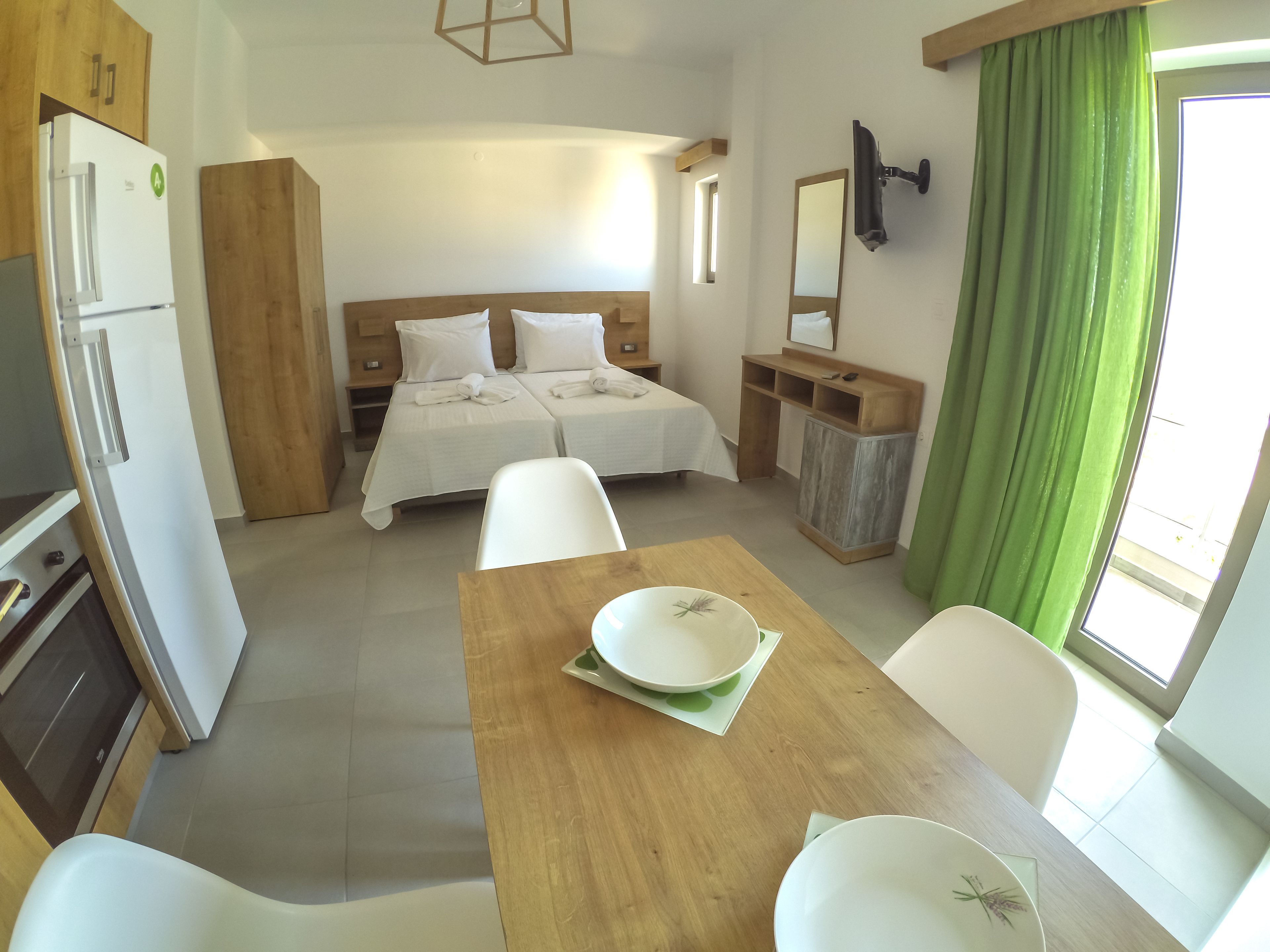 A hotel room with a bed, a desk with chairs, a dining table, a refrigerator, and a green curtain.