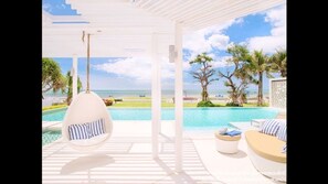 Zona junto a la piscina con una hamaca colgante, un banco acolchado y vistas al océano y palmeras.