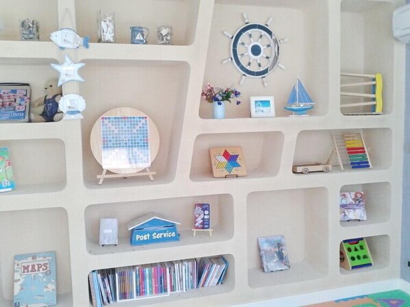 Una habitación con una estantería que contiene varios libros y juguetes, un pequeño velero y un reloj en la pared.