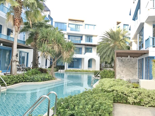 A pool area with a clear blue pool, surrounded by lush greenery and modern buildings with balconies.