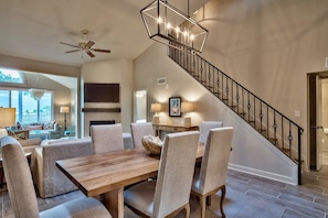A spacious living room with a wooden dining table, comfortable seating, a staircase with a black railing, and a large window offering a view.