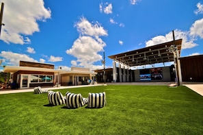 A modern outdoor area with a grassy lawn, striped bean bags, and a covered seating area with a large screen. - Coasting Inn 3 Bedroom Condo By Coastal Dreamin (Santa Rosa Beach)