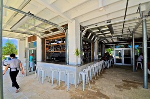 Uma área de bar ao ar livre com um balcão comprido, cadeiras de plástico branco e uma entrada envidraçada.