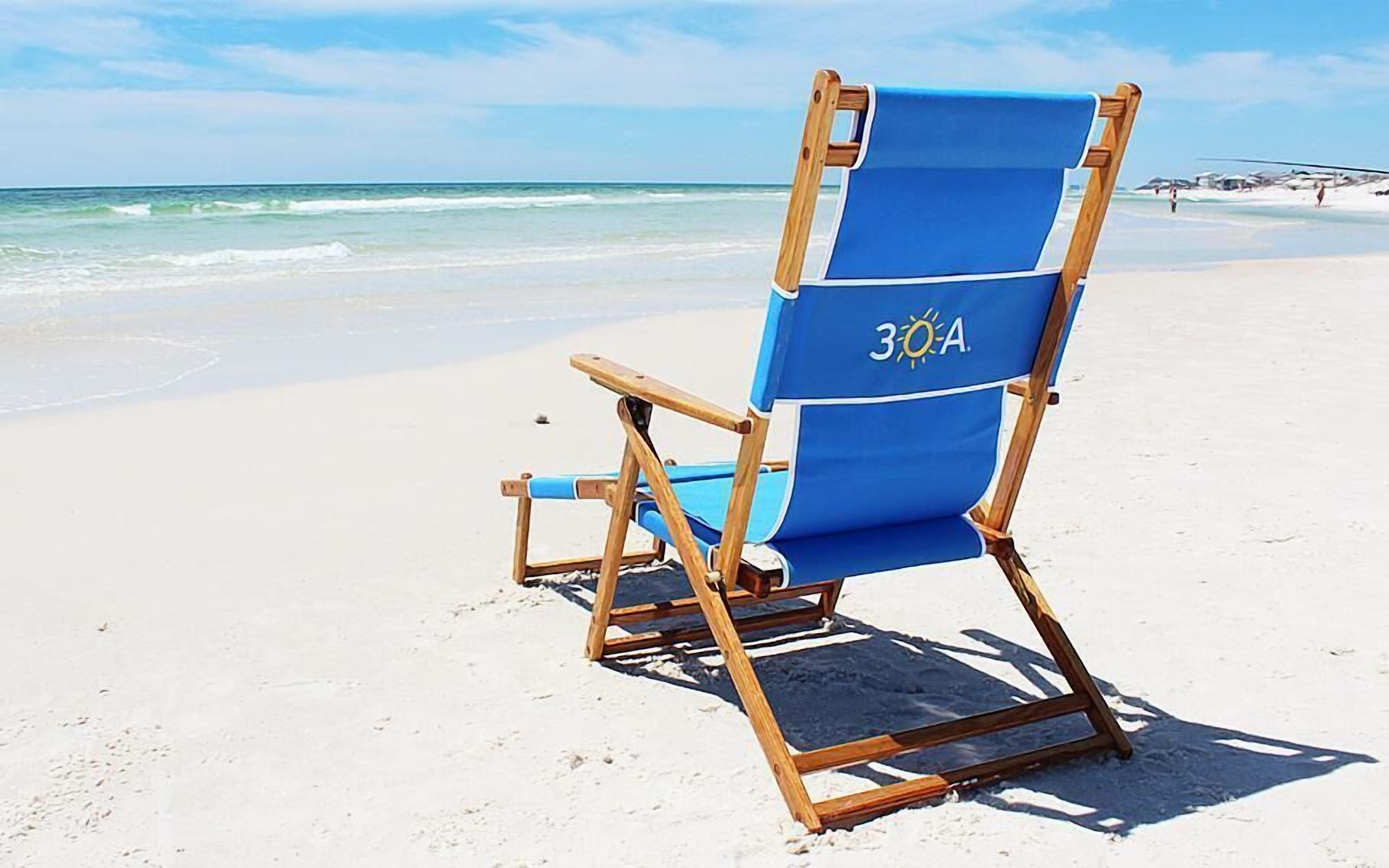 A blue beach chair with a sun logo on the backrest, positioned on a sandy beach with ocean waves in the background.