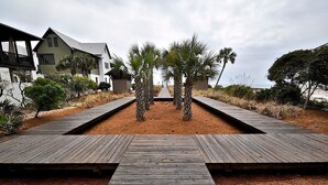 A wooden boardwalk leading to a building with a modern design, surrounded by dry grass and palm trees. - Redfish Village M2 214 3 Bedroom Condo By Coastal Dreamin (Santa Rosa Beach)