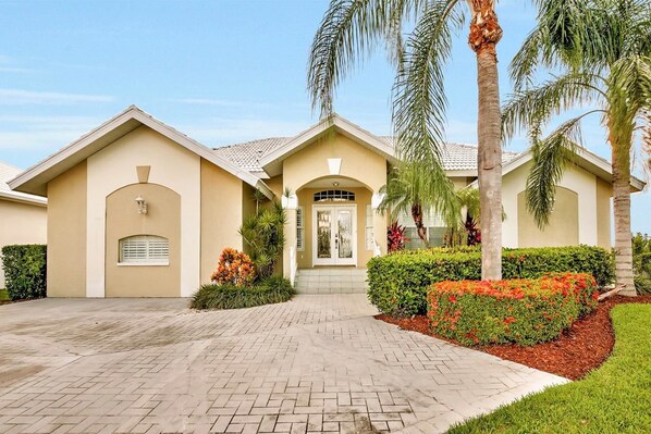 A two-story house with a beige facade, a front entrance with a door and a small window, and a paved driveway. - BE324 4 Bedroom Holiday Home by Marco Naples Vacation Homes (Marco Island)