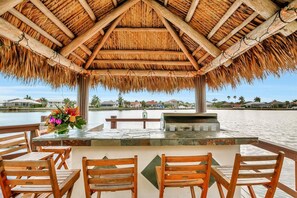 A covered outdoor seating area with wooden chairs and a table, overlooking a body of water with buildings in the background. - BE324 4 Bedroom Holiday Home by Marco Naples Vacation Homes (Marco Island)