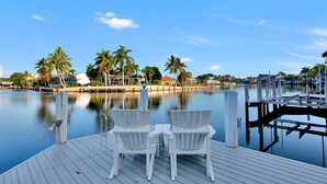 A wooden dock with two white chairs facing a calm body of water, surrounded by palm trees and buildings in the background. - COPPER20 5 Bedroom Holiday Home by Marco Naples Vacation Homes (Marco Island)