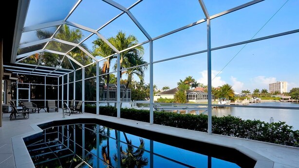 A modern indoor pool with a glass ceiling, surrounded by lounge chairs and a view of a body of water and palm trees. - COPPER20 5 Bedroom Holiday Home by Marco Naples Vacation Homes (Marco Island)