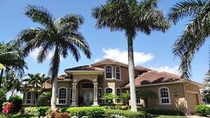 Una casa grande de dos pisos con fachada marrón y techo de tejas, rodeada de césped cuidado y palmeras.