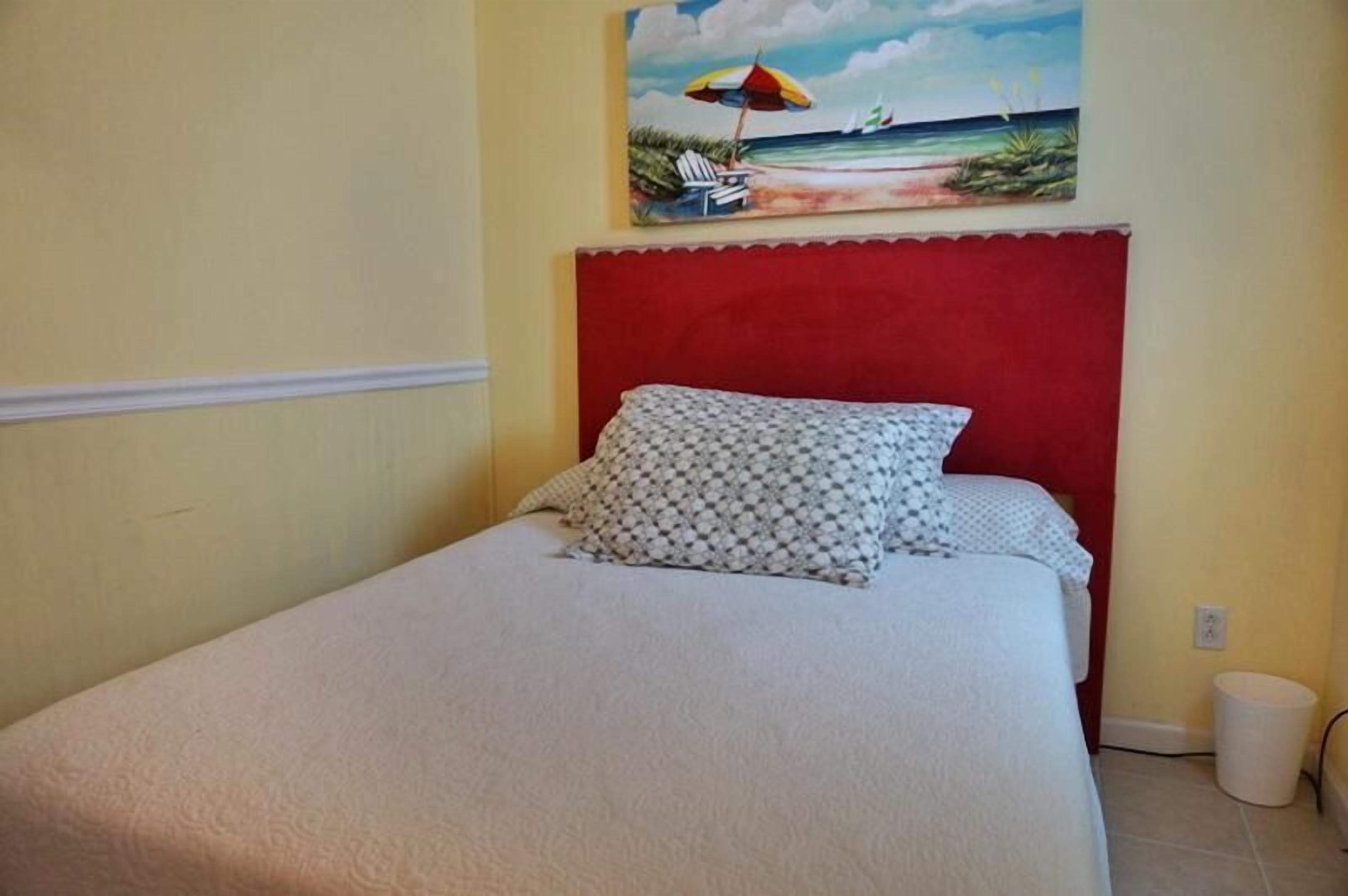 A single bed with a red headboard, a patterned pillow, and a beach scene painting above it.