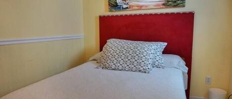 A single bed with a red headboard, a patterned pillow, and a beach scene painting above it.