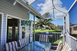 Un espace extérieur aménagé, abrité et doté d’une table en verre et de coussins à rayures, offrant une vue sur une piscine et des palmiers.