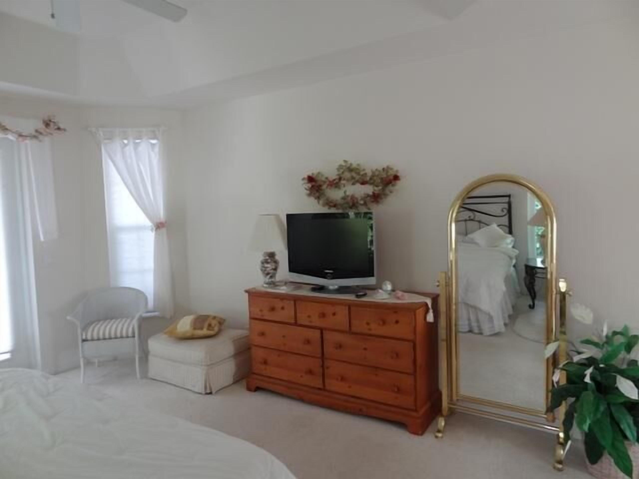 Een slaapkamer met een bed, een dressoir, een televisie, een stoel, een spiegel en een plant.
