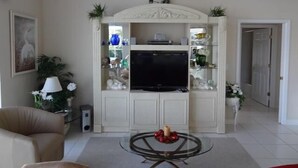 A living room with a beige sofa, a glass coffee table, a flat-screen TV, and a decorative shelf with various items. - KEN486 3 Bedroom Holiday Home by Marco Naples Vacation Homes (Marco Island)