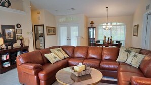 A living room with a brown leather sectional sofa, a glass coffee table, and a dining area with a chandelier.