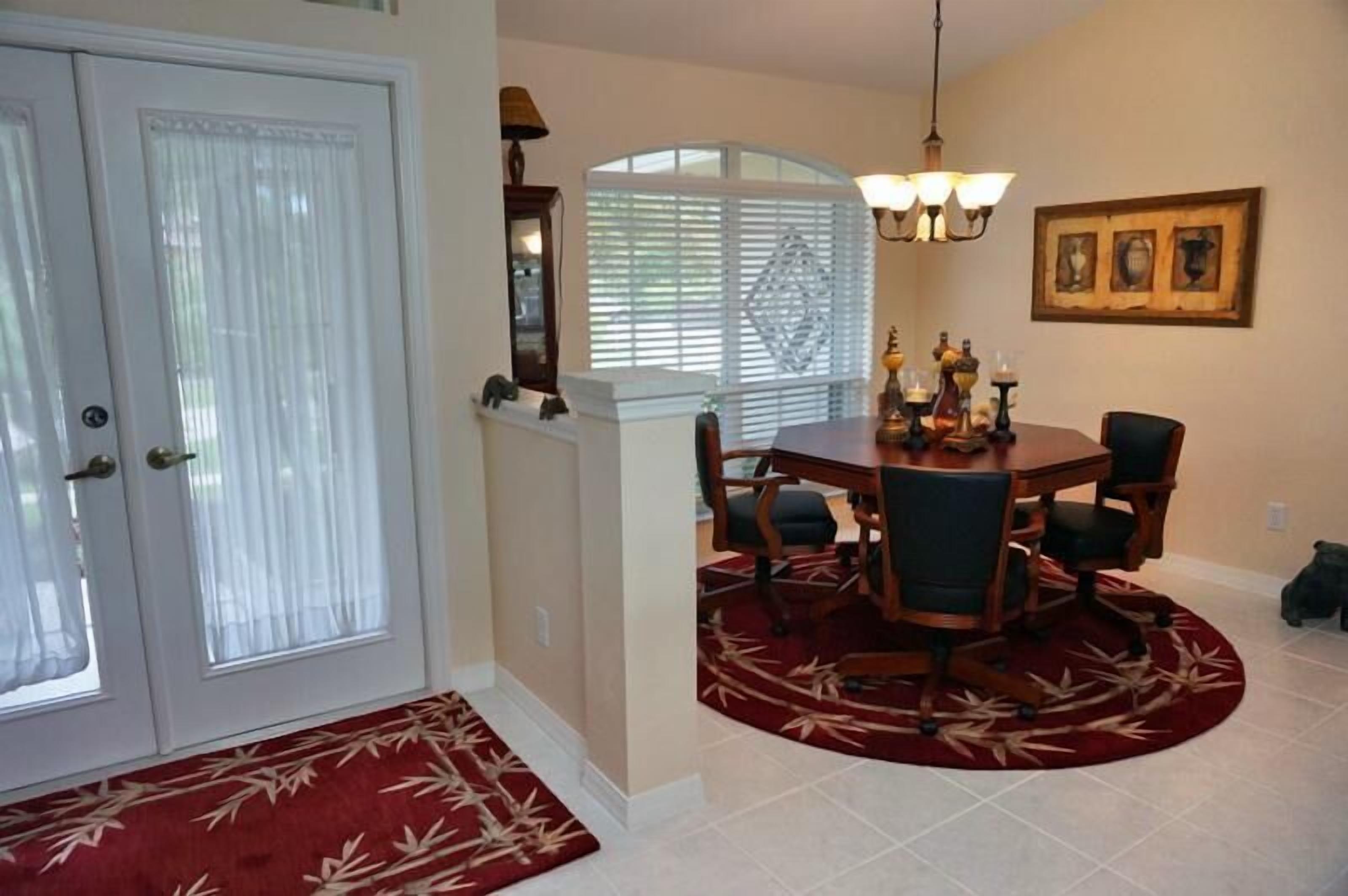 A dining area with a round table, chairs, a red patterned rug, a chandelier, and a window with blinds.