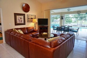 Ein Wohnzimmer mit braunem Ledersofa, einem Fernseher und einem Tisch. Es gibt eine Terrasse mit Schwimmbad und Stühlen.