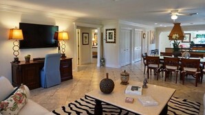 A spacious living room with a flat-screen TV, a coffee table with decorative items, a dining area with wooden chairs, and a ceiling fan.