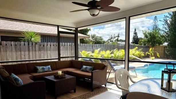 A sunlit patio with a sofa, chairs, and a small table, overlooking a swimming pool and lush greenery. - NP97TH 730 3 Bedroom Holiday Home by Marco Naples Vacation Homes (Naples)