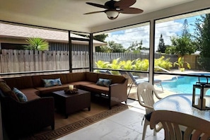 A sunlit patio with a sofa, chairs, and a small table, overlooking a swimming pool and lush greenery.