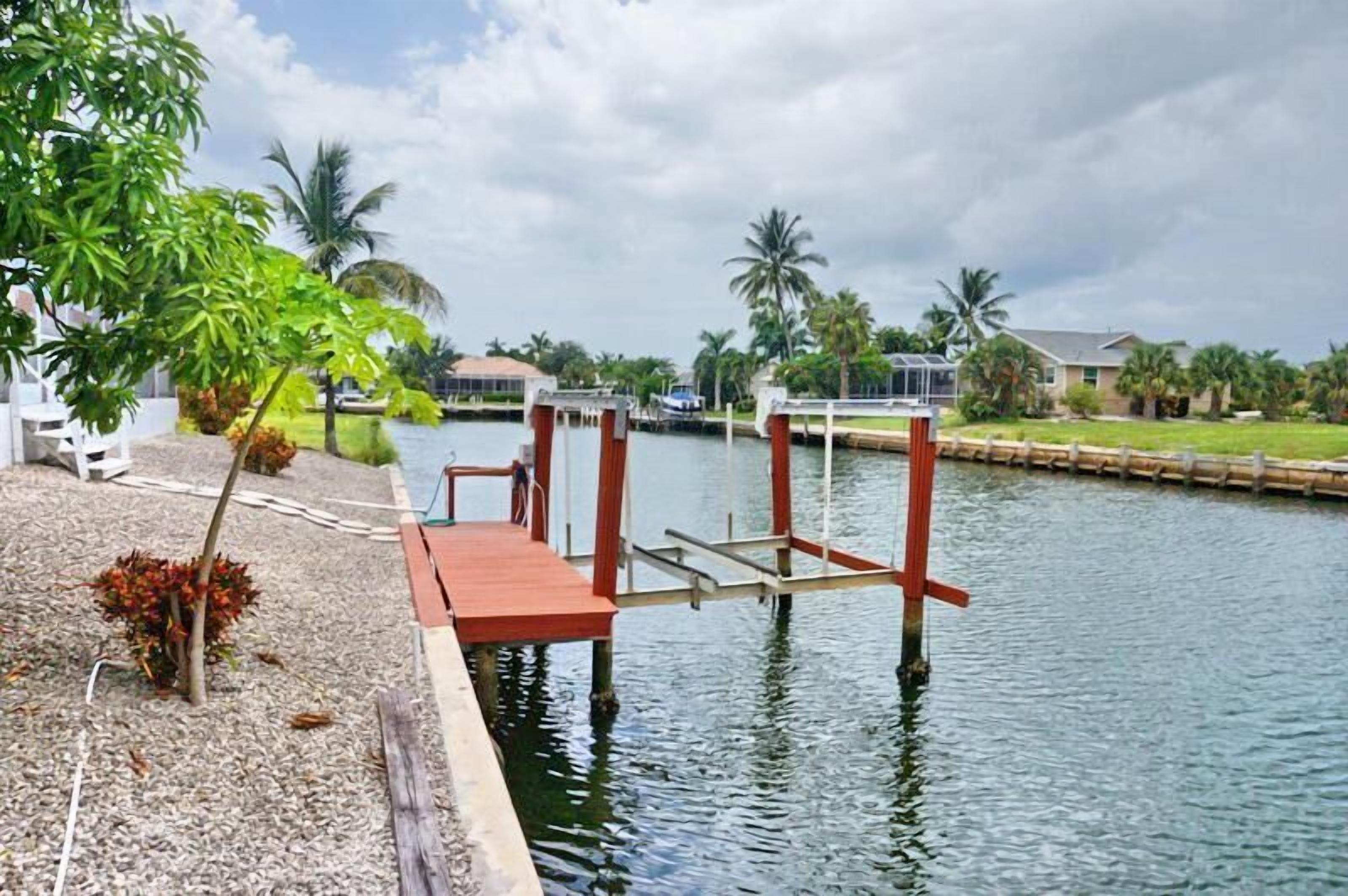 A dock extending into calm water with a clear sky, surrounded by palm trees and residential buildings.