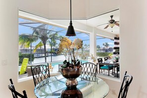Ein Essbereich mit einem Glastisch, Stühlen, einer dekorativen Schale mit Blumen und Blick auf einen Pool und Palmen.