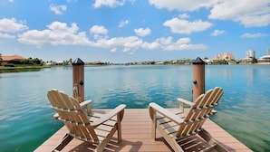 A wooden deck with two Adirondack chairs facing a calm lake, surrounded by buildings and a clear sky.