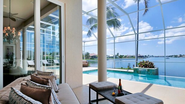 A modern living room with a large glass door leading to a pool area, featuring a sofa, coffee table, and a view of the water.