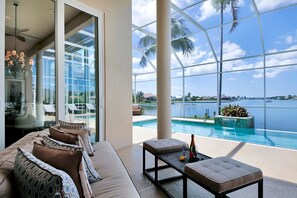 A modern living room with a large glass door leading to a pool area, featuring a sofa, coffee table, and a view of the water. - SMARCO919 5 Bedroom Holiday Home by Marco Naples Vacation Homes (Marco Island)