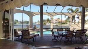 A covered outdoor area with a pool, seating, and a view of a body of water and palm trees.