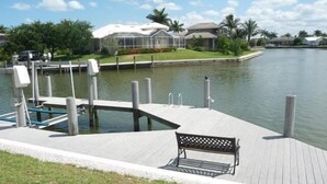 Un muelle frente al agua con un banco, piscina y casas al fondo.