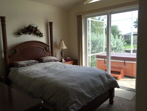 Un dormitorio con una cama grande, una mesita de noche con lámpara, una ventana con vista a follaje, y un cabecero de madera.