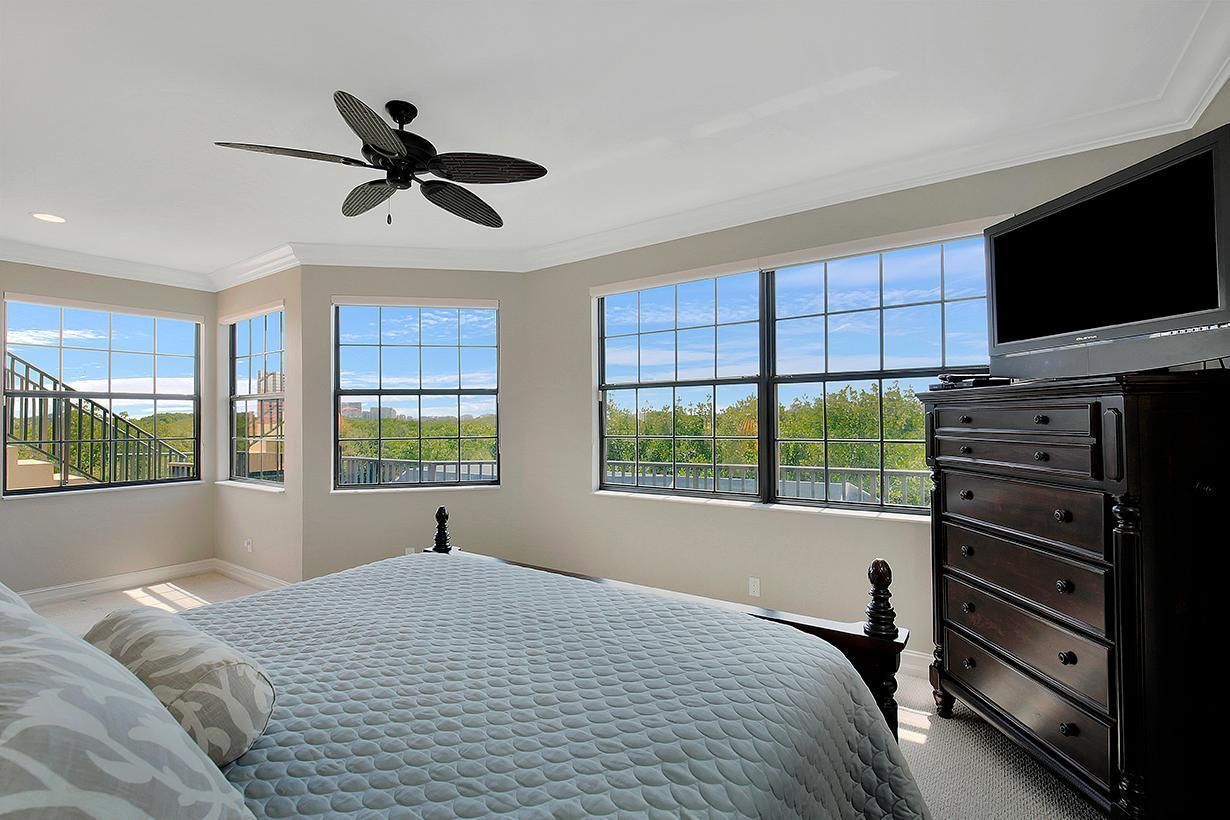 A bedroom with a large bed, a ceiling fan, a dresser, and a TV. There are large windows with a view of greenery and a staircase.