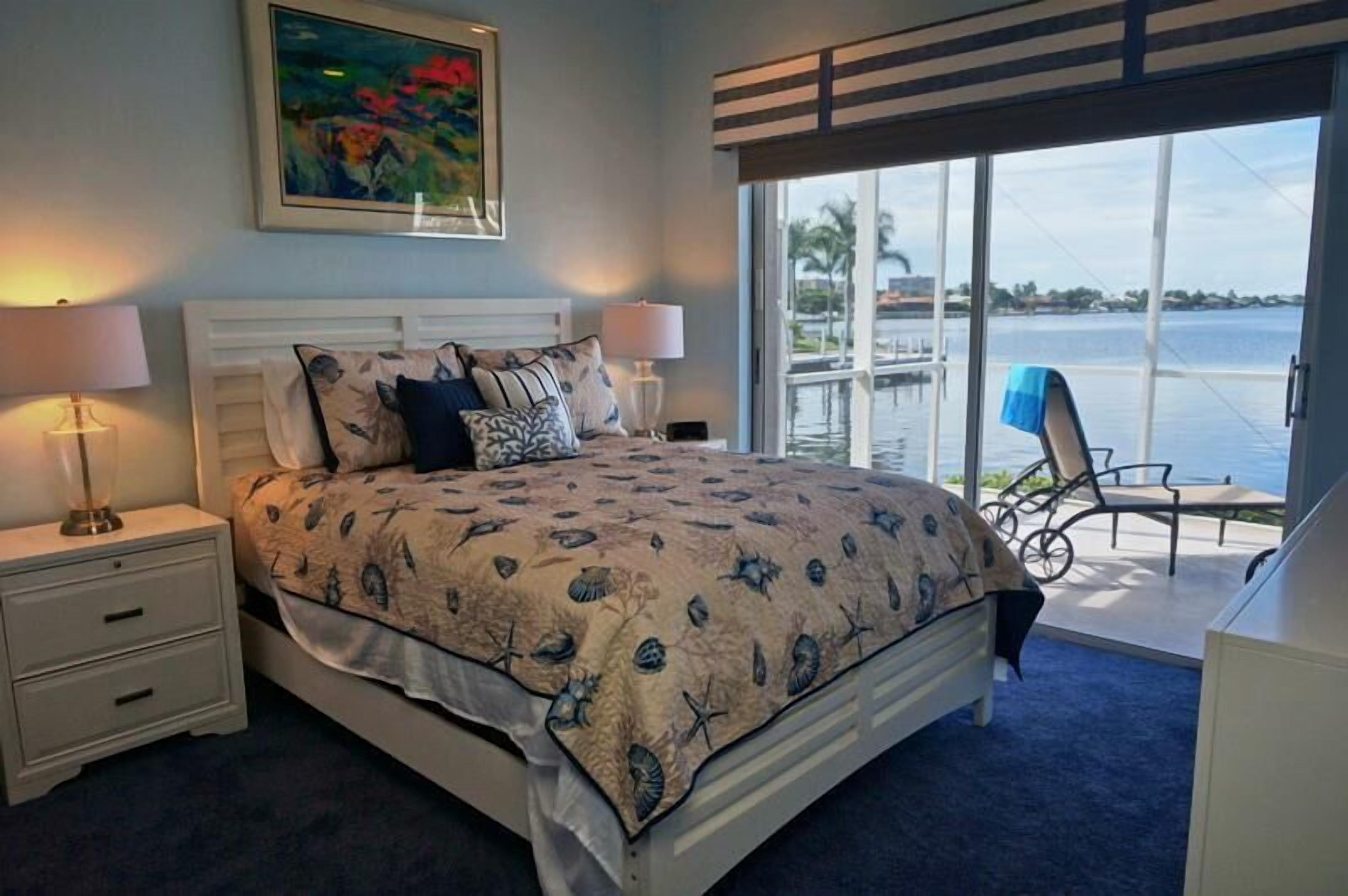 Ein Schlafzimmer mit einem Bett, zwei Nachttischlampen, einem Nachttisch, einem Sessel und Blick auf das Wasser durch große Fenster.