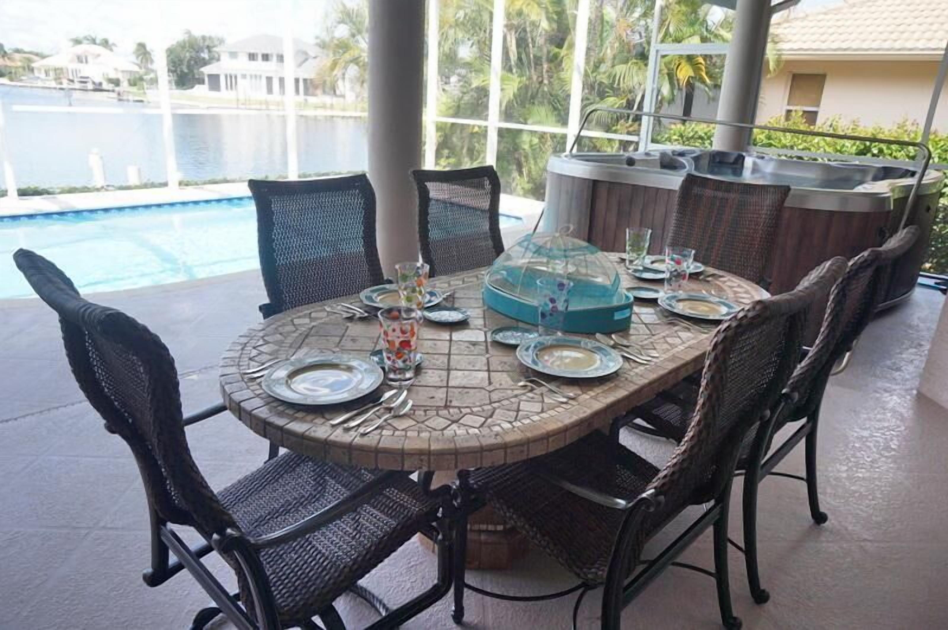 An outdoor dining area with a round table set for six, surrounded by wicker chairs. A hot tub is visible in the background, and the setting overlooks a body of water.