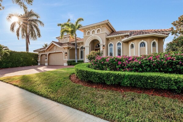 A large, single-story house with a beige facade, a tiled roof, and a well-manicured front yard with a hedge and flowering plants.