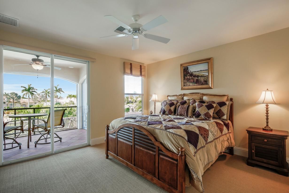 A bedroom with a large bed, a ceiling fan, a painting, and a view of palm trees through a sliding door.