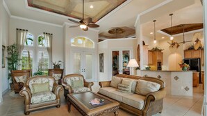 A spacious living room with a sofa, armchairs, and a coffee table. There is a kitchen area with cabinets and a refrigerator.