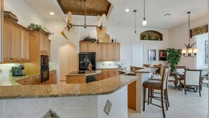 A spacious kitchen with granite countertops, wooden cabinets, and a central island. There is a dining area with wooden chairs and a chandelier.