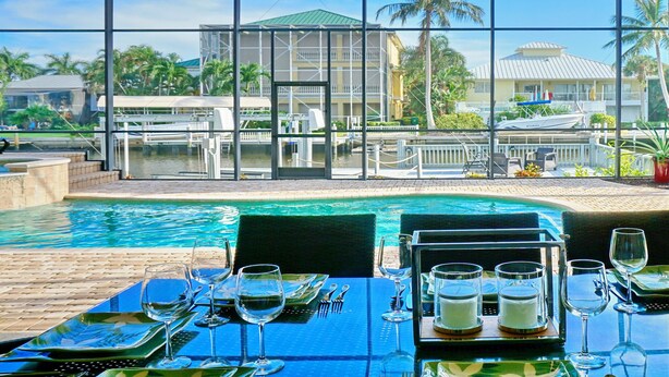 Uma área de refeições com mesas preparadas para uma refeição, com vista para uma piscina e para um edifício com telhado verde e palmeiras.