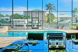 A dining area with tables set for a meal, overlooking a swimming pool and a view of a building with a green roof and palm trees.