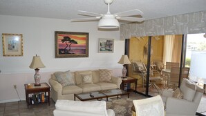 A living room with a sofa, coffee table, and chairs. There are bookshelves, a lamp, and artwork on the walls.