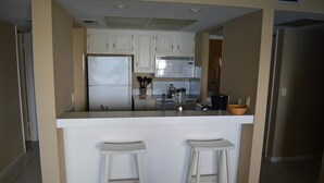 A kitchen with white cabinets, a stainless steel refrigerator, a microwave, and a blender on the counter. There are two white bar stools. - Cove II 127F 2 Bedroom Condo By Midnight Cove II (Siesta Key)
