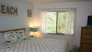 A bedroom with a bed, a bedside lamp, a window with curtains, a television on a wooden cabinet, and a framed picture on the wall. - Cove II 138F 2 Bedroom Condo By Midnight Cove II (Siesta Key)