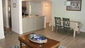 A compact kitchen with white cabinets, a microwave, and a small island. There is a dining area with a table and chairs, and a wooden coffee table with a tray. - Cove II 138F 2 Bedroom Condo By Midnight Cove II (Siesta Key)