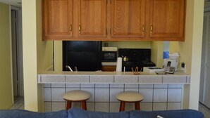 A kitchen with wooden cabinets, a black refrigerator, an oven, a coffee maker, and two bar stools. - Cove II 233F 2 Bedroom Condo By Midnight Cove II (Siesta Key)